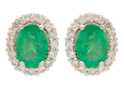 Lot 169 - A pair of 18ct white gold oval emerald and diamond set cluster stud earrings.